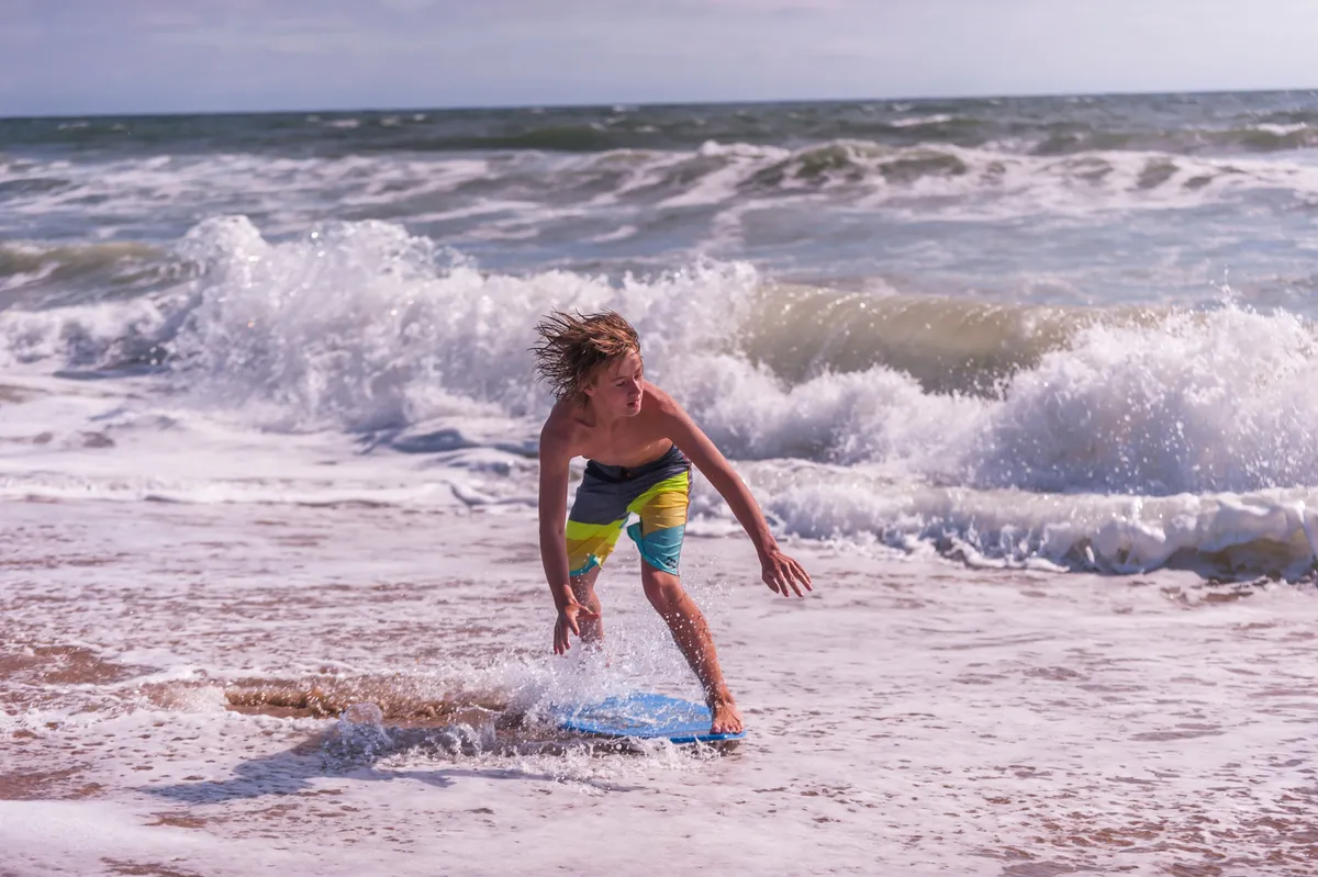 A joyful, sunlit moment featuring a boy riding a boogie board in the surf, with energetic waves and children playing in the sand nearby. The scene feels lively and adventurous, emphasizing fun water activities and the thrill of the ocean.