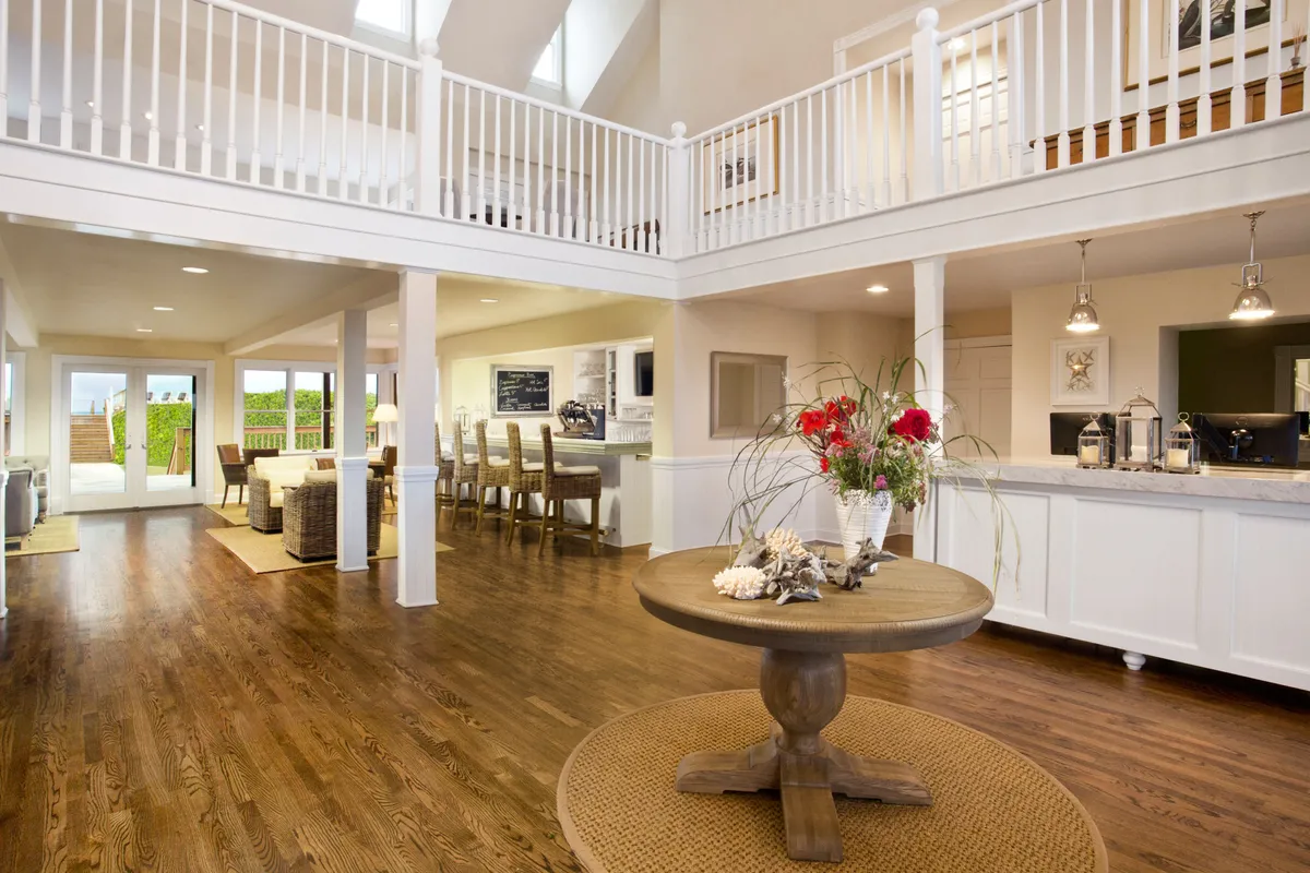 A spacious, light-filled open-plan area featuring a round dining table in the foreground, an open kitchen in the background, and large windows allowing natural light to stream in. The design conveys an inviting, accessible space suitable for gatherings or relaxed moments, with wide pathways and uncluttered layouts that support ease of movement.
