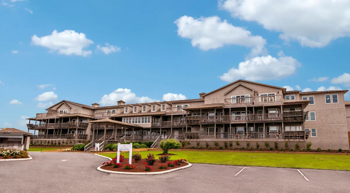 This image features the building's facade with several balconies, each equipped with seating that overlooks the coastline. The landscaping is lush and inviting, while subtle signage and people relaxing on the balconies evoke a sense of peaceful leisure.