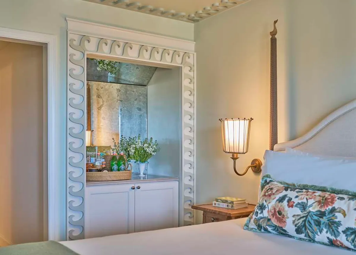 A tranquil guestroom scene featuring a decorative mirror above a bedside table, a cozy lamp casting a soft glow, a floral cushion atop plush bedding, and a touch of greenery in a vase. The large windows hint at resort views and allow natural light to fill the space, emphasizing comfort and understated elegance.