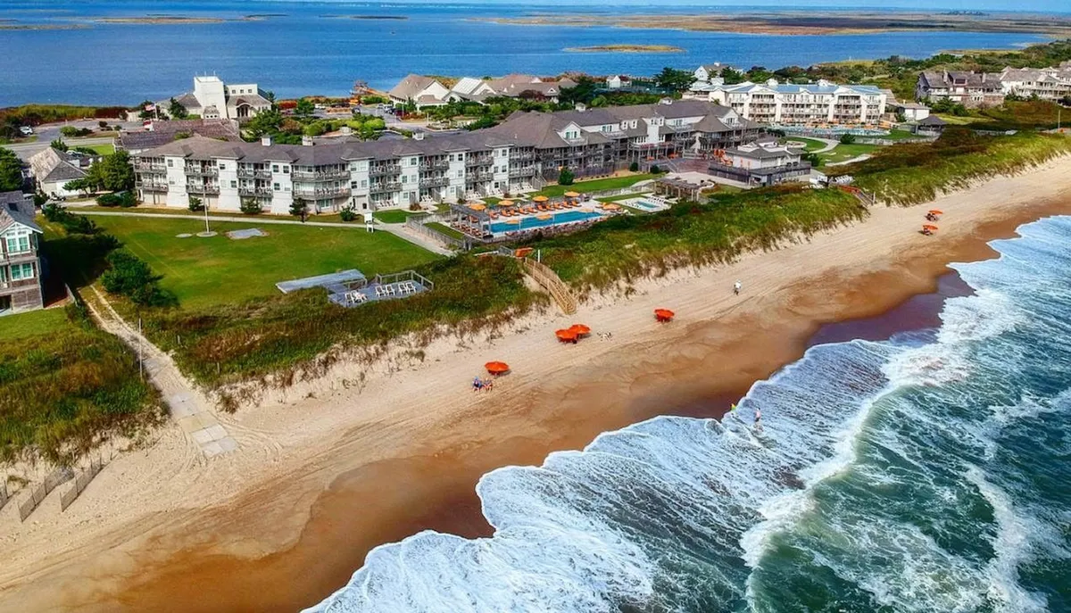 A wide-angle view of the beachfront property, showcasing both the swimming pool with orange umbrellas and the ocean waves crashing nearby. The scene exudes a carefree vacation vibe, with spaces for sunbathing, swimming, and family games.