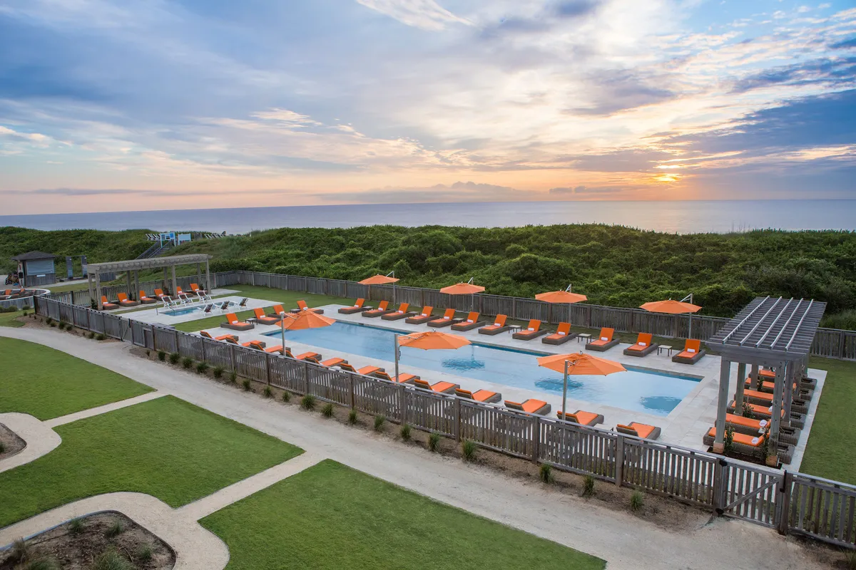 A lively resort pool area surrounded by orange umbrellas and comfortable lounge chairs, with guests enjoying swimming and socializing. In the distance, the sun is setting over the ocean, creating a memorable and vibrant backdrop for family activities.