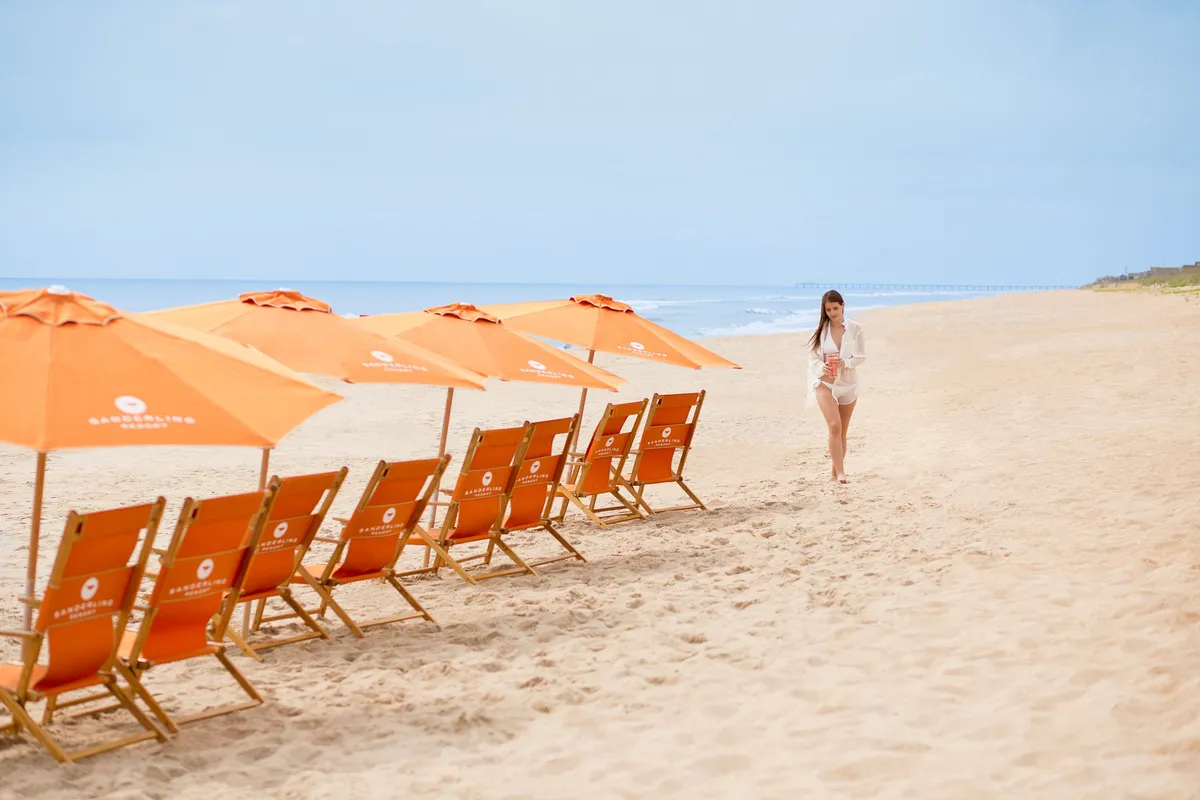 This image captures a serene beach setting with orange beach chairs and umbrellas arranged in the sand, overlooking the ocean. A woman is seen walking along the shoreline, evoking a peaceful, leisurely atmosphere. The scene highlights private, comfortable relaxation spaces directly on the beach, with the ocean stretching out beyond.