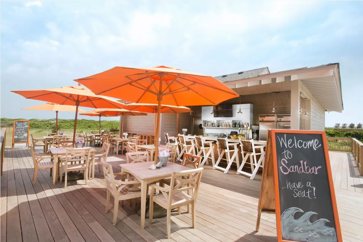 An outdoor dining area featuring wooden tables under cheerful orange umbrellas, with the Sandbar structure and a welcome sign clearly visible. The setup evokes a relaxed, breezy vibe ideal for outdoor dining, drinks, and live music. The background hints at open-air enjoyment and coastal ambiance.