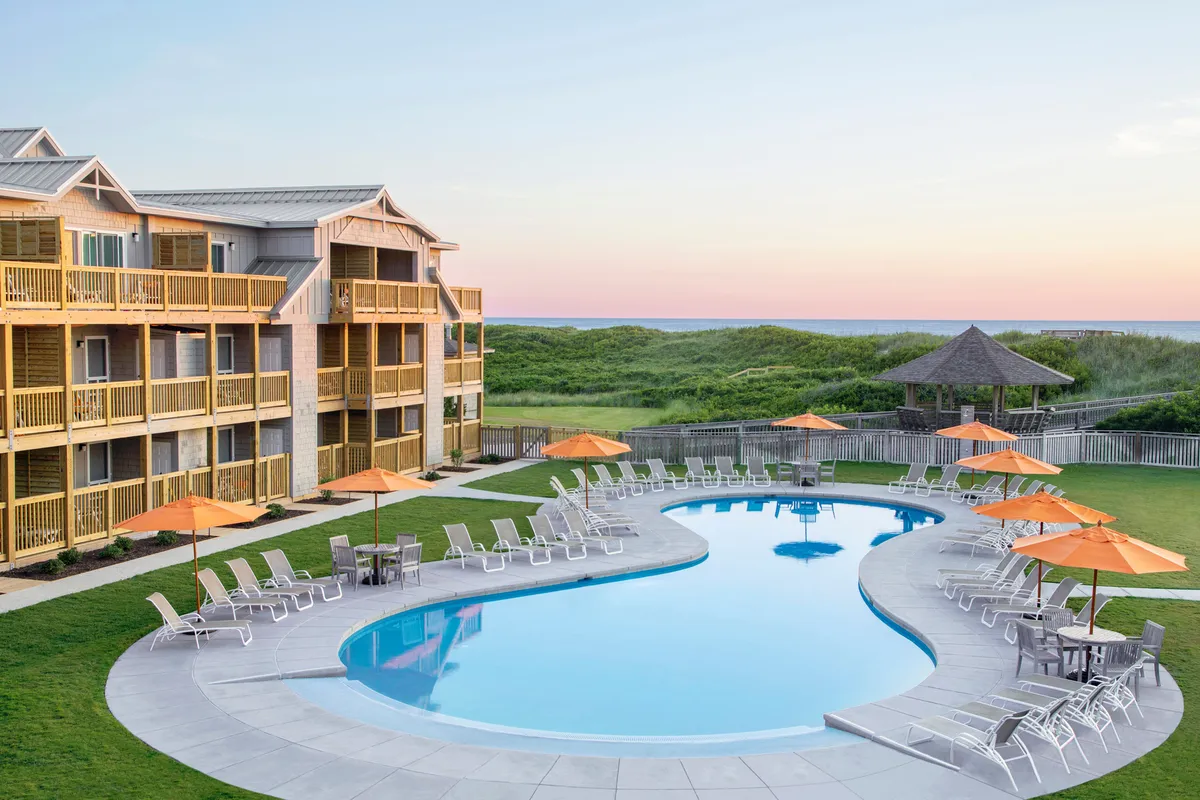 A wide-angle exterior view of the main swimming pool area, with elegant lounge chairs and umbrellas arranged around the water. The hotel facade is visible in the background, and the lighting suggests either early morning or sunset, enhancing the tranquil and inviting mood. Groups of guests are seen swimming, sunbathing, or relaxing by the pool, contributing to a communal and restful setting.