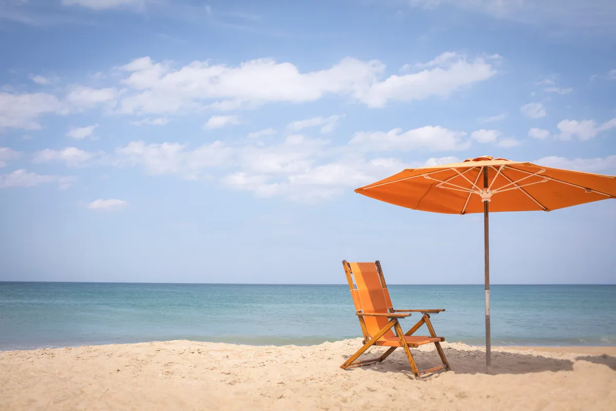 A tranquil stretch of private beach featuring an inviting orange beach chair and umbrella set up on the sand, with gentle waves lapping in the background. The scene exudes relaxation and accessibility, suggesting a perfect spot for families to unwind and play just steps from their accommodations.