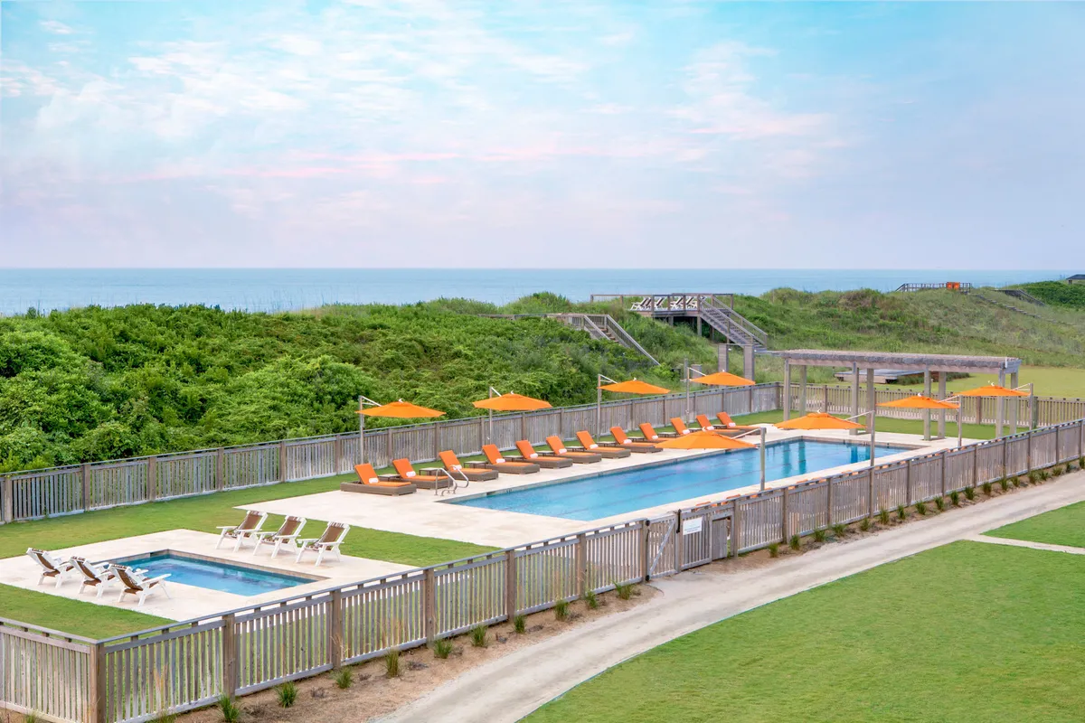 This image captures a spacious outdoor swimming pool surrounded by comfortable lounge chairs and vivid orange umbrellas, set against a lush backdrop of landscape greenery. The scene includes guests enjoying the pool, socializing, and relaxing in the sun, illustrating a lively yet leisurely atmosphere.