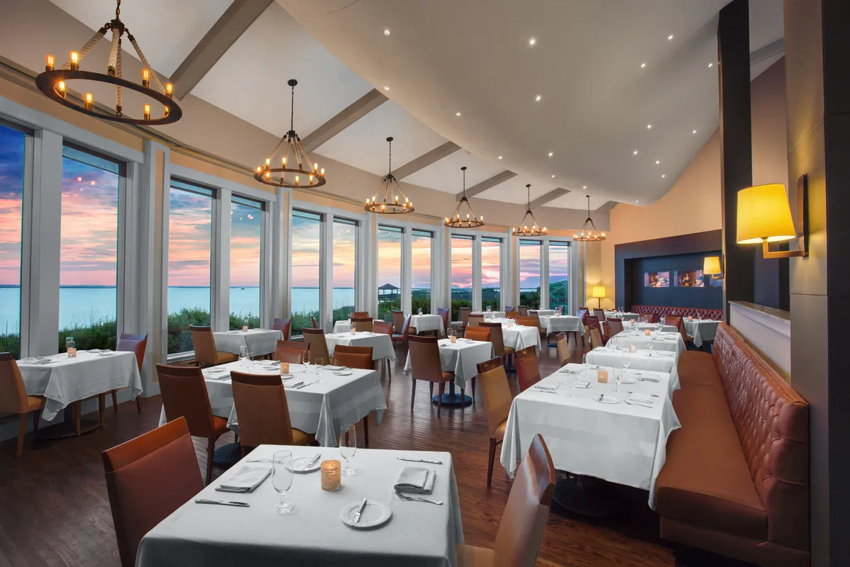 This image shows a stylish dining room with large windows framing a beautiful sunset view. Elegant chandeliers hang above neatly arranged tables, setting the scene for refined dining experiences. The room exudes a warm, inviting glow, emphasizing both the culinary sophistication and the social ambiance.