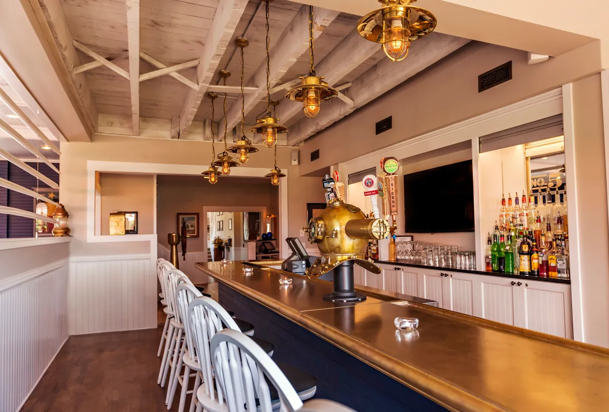 The image captures a lively, brass-accented bar area. Rows of bottled liquors, hanging lights, and decorative touches create a relaxed yet sophisticated vibe, perfect for socializing, enjoying cocktails, or casual dining. The inviting bar counter suggests a friendly, bustling environment for guests to gather.
