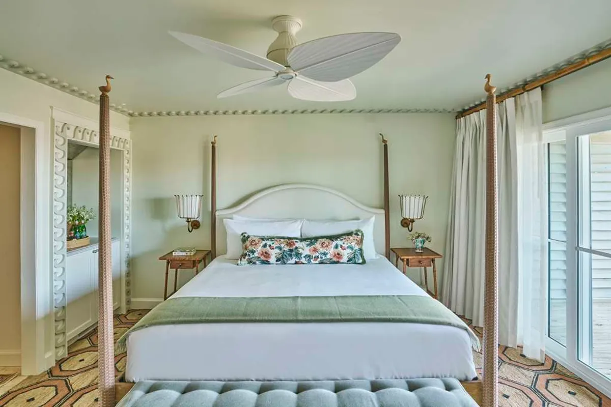 The image showcases a serene studio interior with a four-poster bed draped in luxury bedding and decorative pillows, complemented by a mirrored dresser and a ceiling fan overhead. Sunlight gently streams into the room, creating a peaceful atmosphere perfect for relaxation, reading, or meditation.